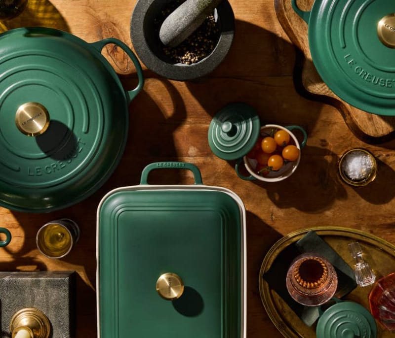 A styled flat-lay of Le Creuset cookware in a deep forest green enamel finish with polished brass handles and knobs, arranged on a rustic wooden table alongside spices, a mortar and pestle, and serving dishes.